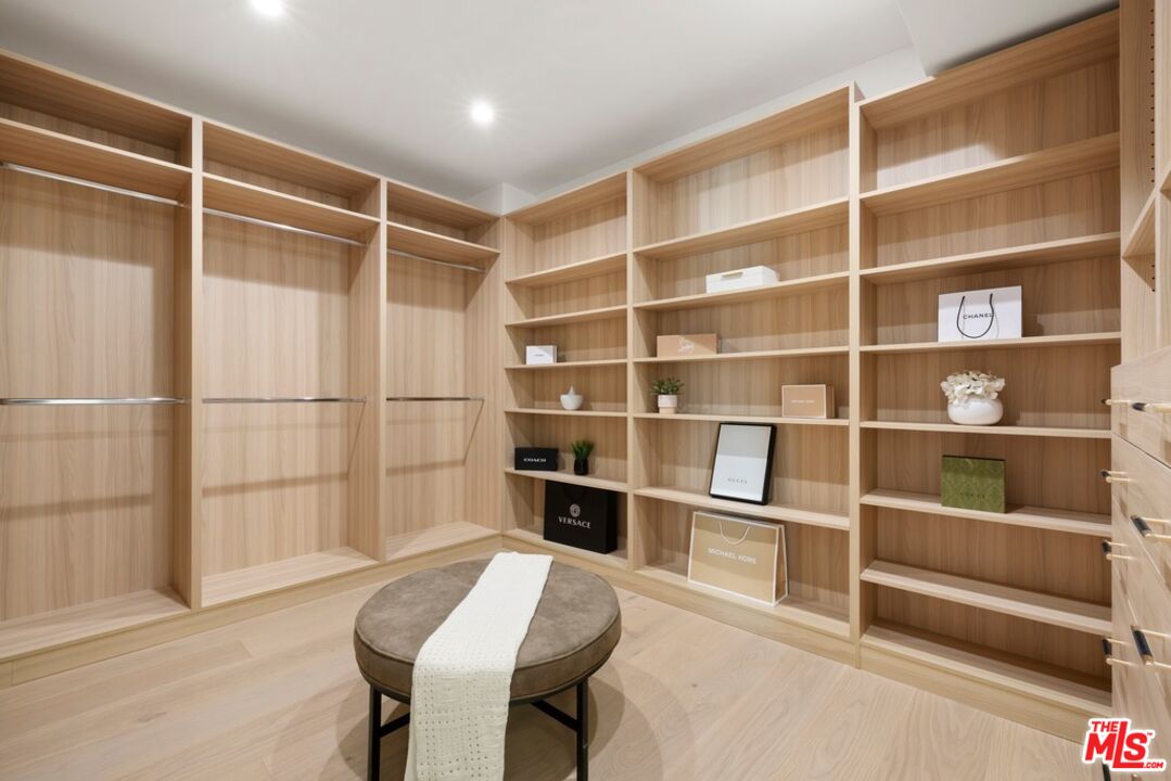 Walk-in closet with light wood shelving, hanging rails, and a round ottoman with a white throw.