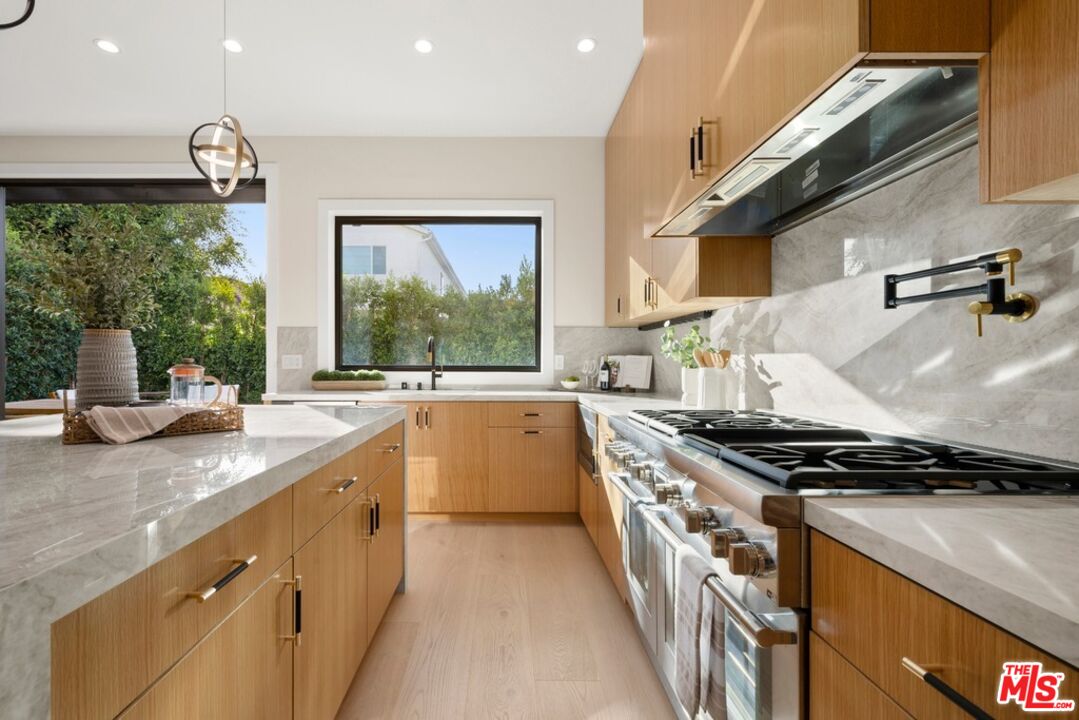 Modern kitchen with oak cabinets, marble counters, and a large gas range beside a garden-view window.