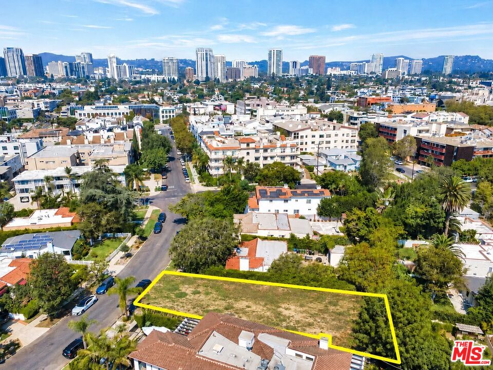 Aerial view of a vacant lot outlined in yellow among homes, trees, and a distant city skyline.