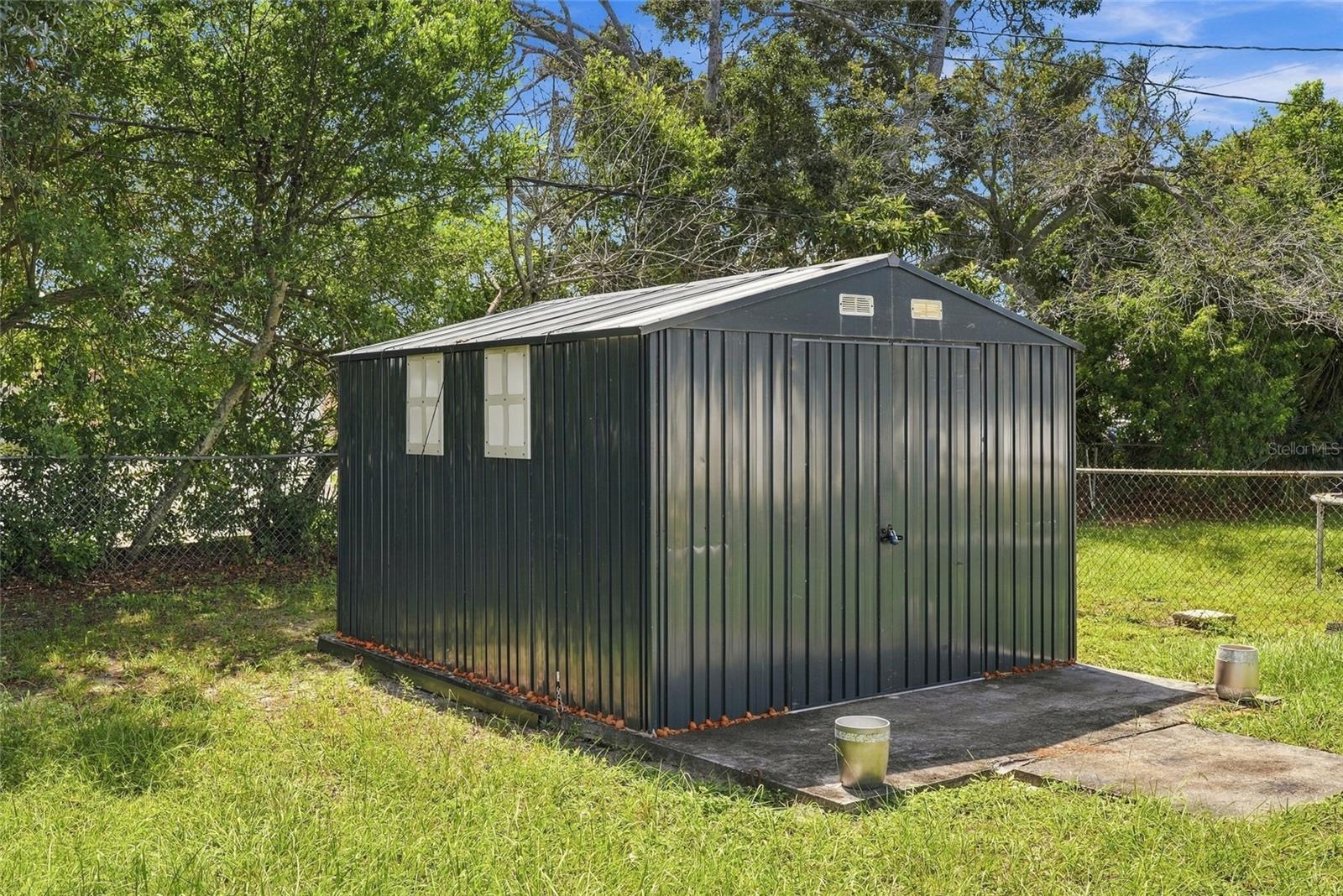 Dark metal storage shed on a concrete pad in a grassy, tree-lined yard.