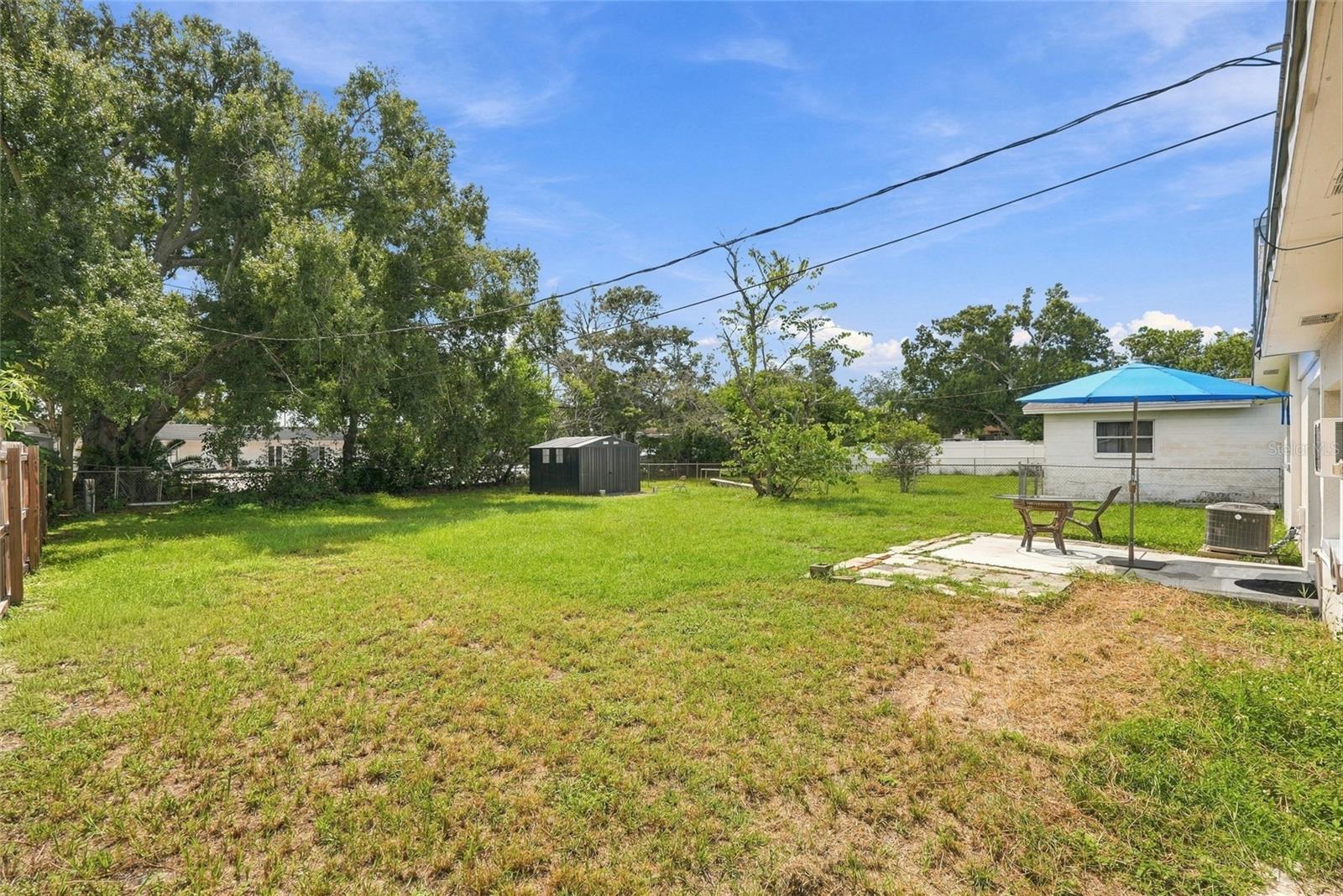 Fenced grassy backyard with a patio table under a blue umbrella and a dark storage shed.