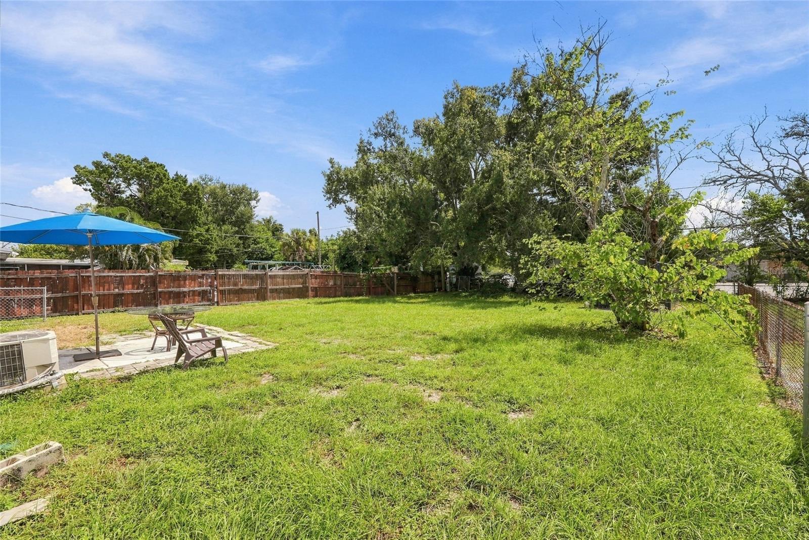 Fenced grassy backyard with a paved patio, seating, blue umbrella, and mature trees.