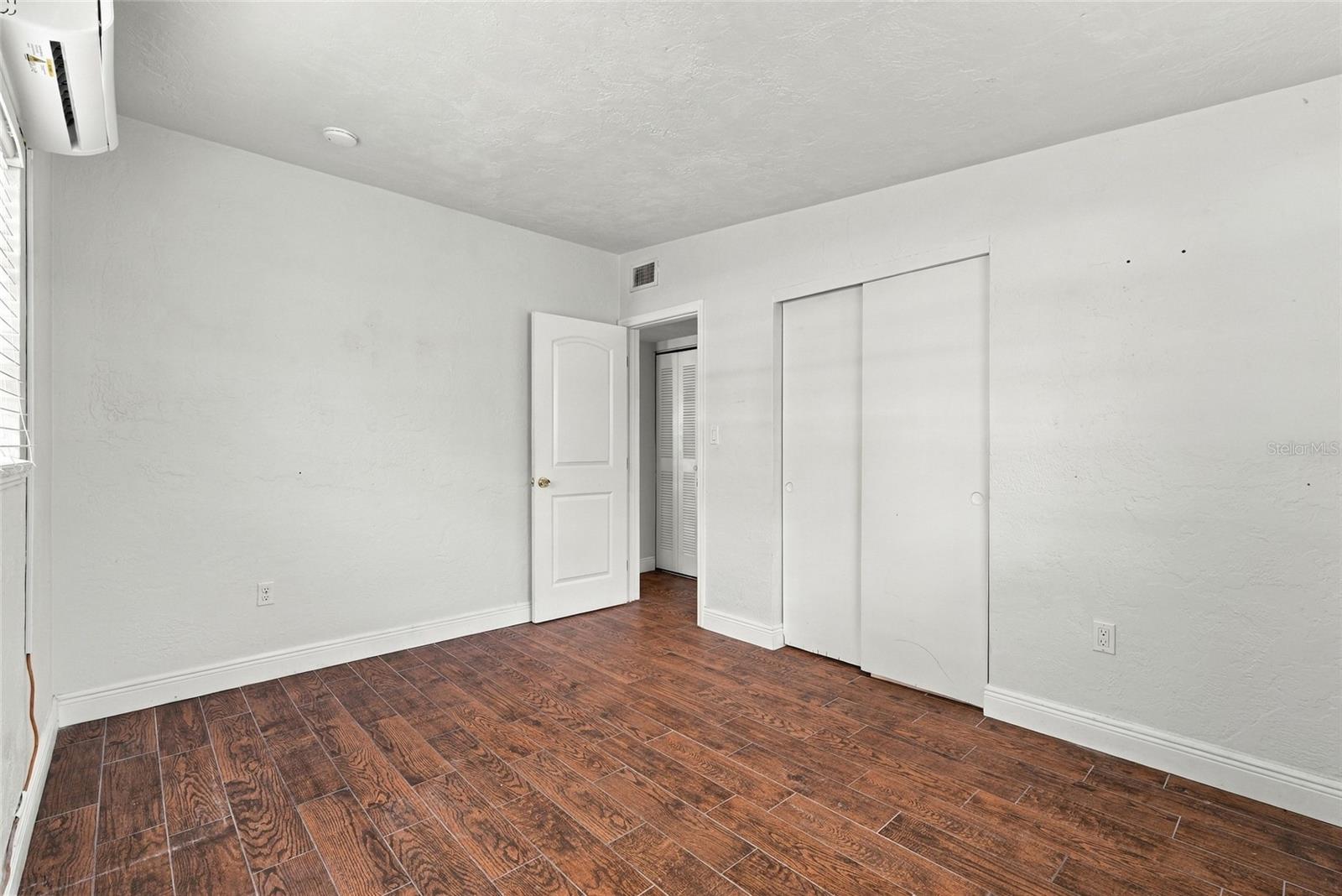 Empty bedroom with reddish-brown wood-look tile flooring, sliding closet doors, and a wall-mounted air conditioner.