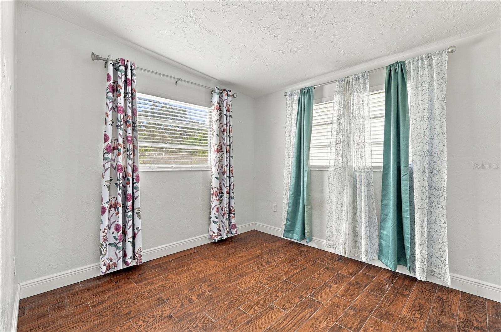 Empty bedroom with wood-look flooring and two curtained windows.