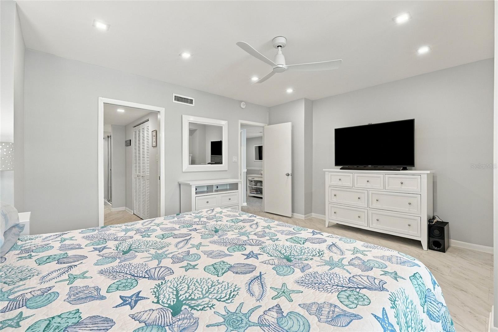 Bedroom with a seashell-patterned quilt, white dresser, wall-mounted television, and ceiling fan.