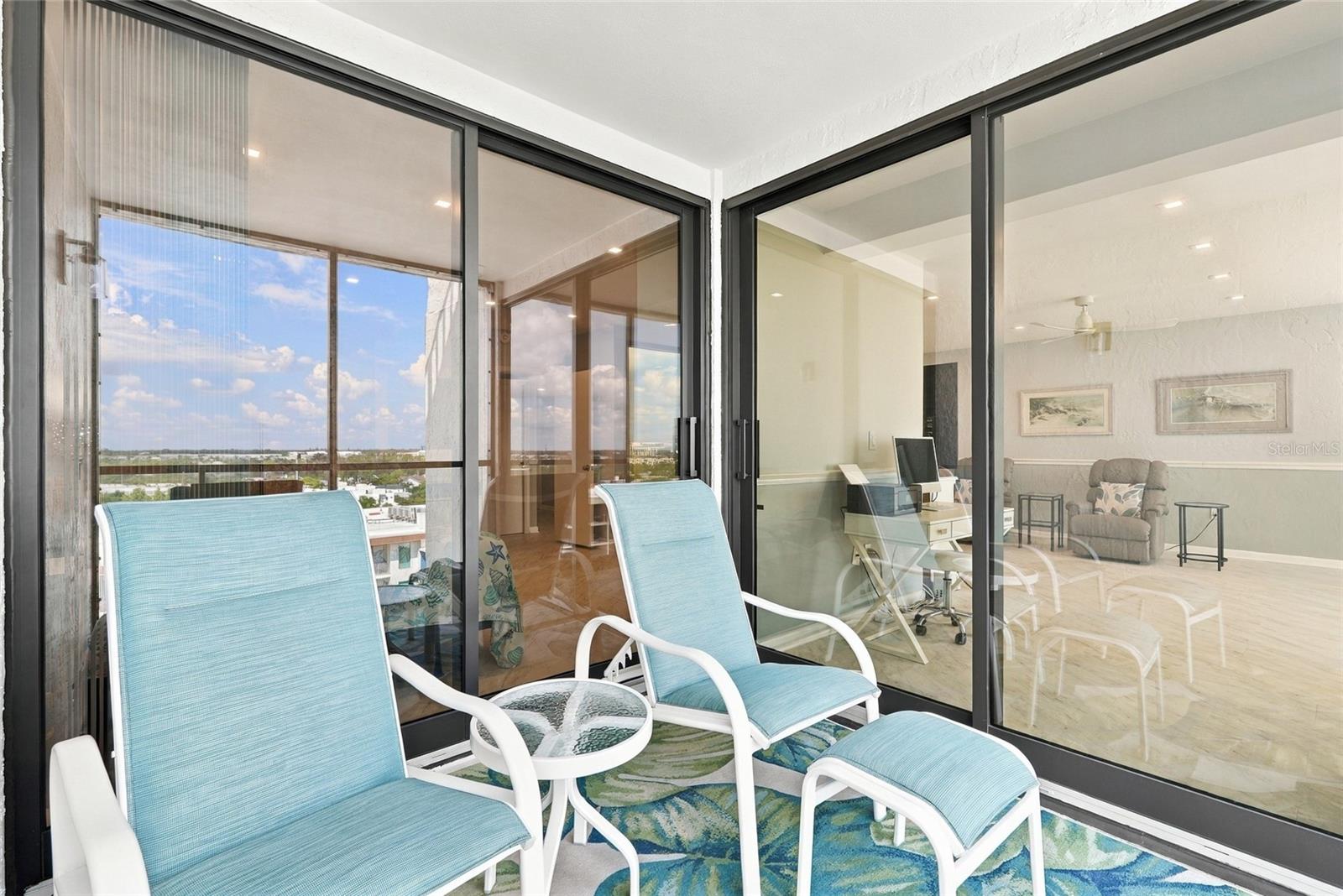 Condo balcony with blue lounge chairs, glass doors, and an elevated view of nearby buildings.