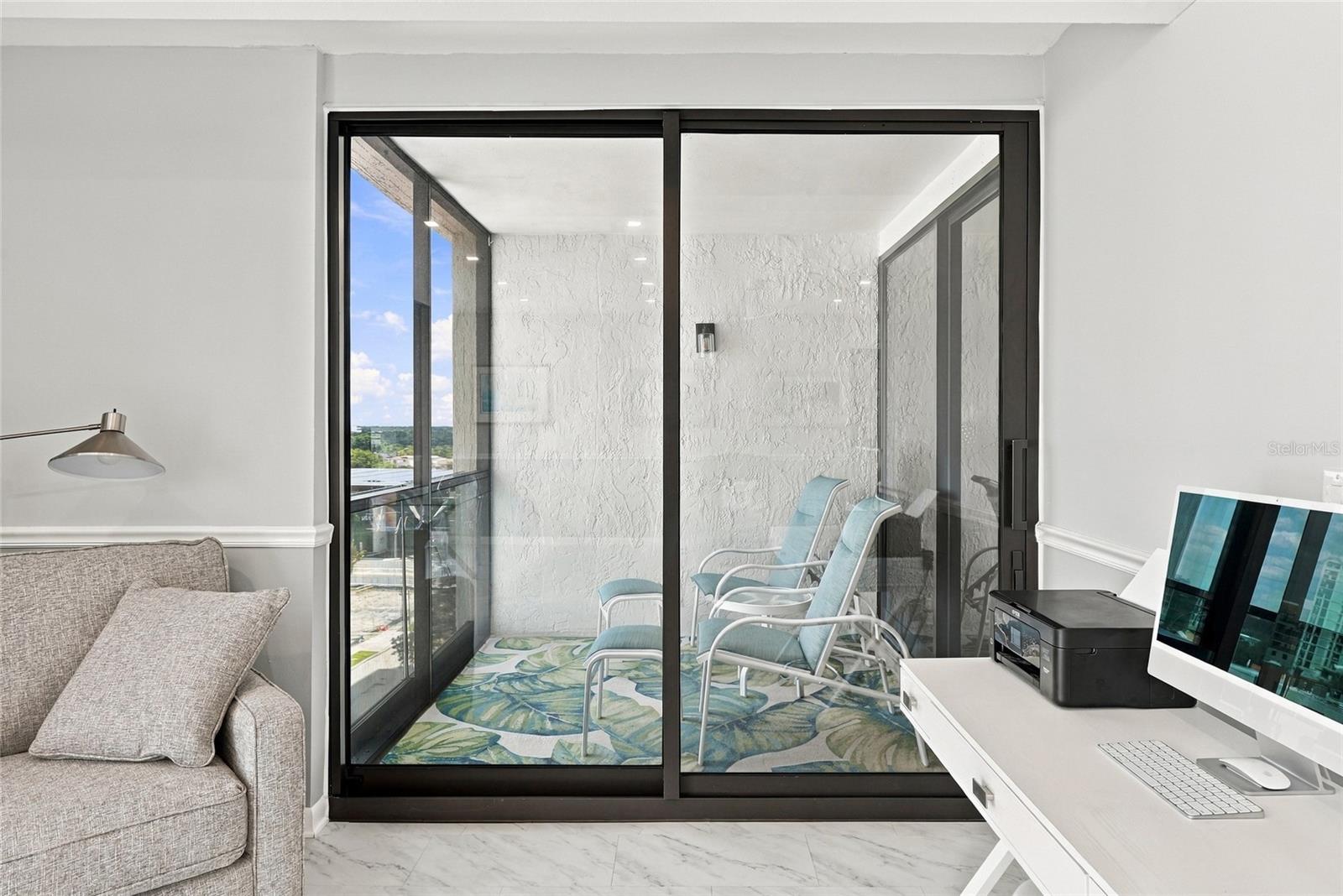 Condo living room with a gray sofa and desk overlooking a covered balcony with chairs.