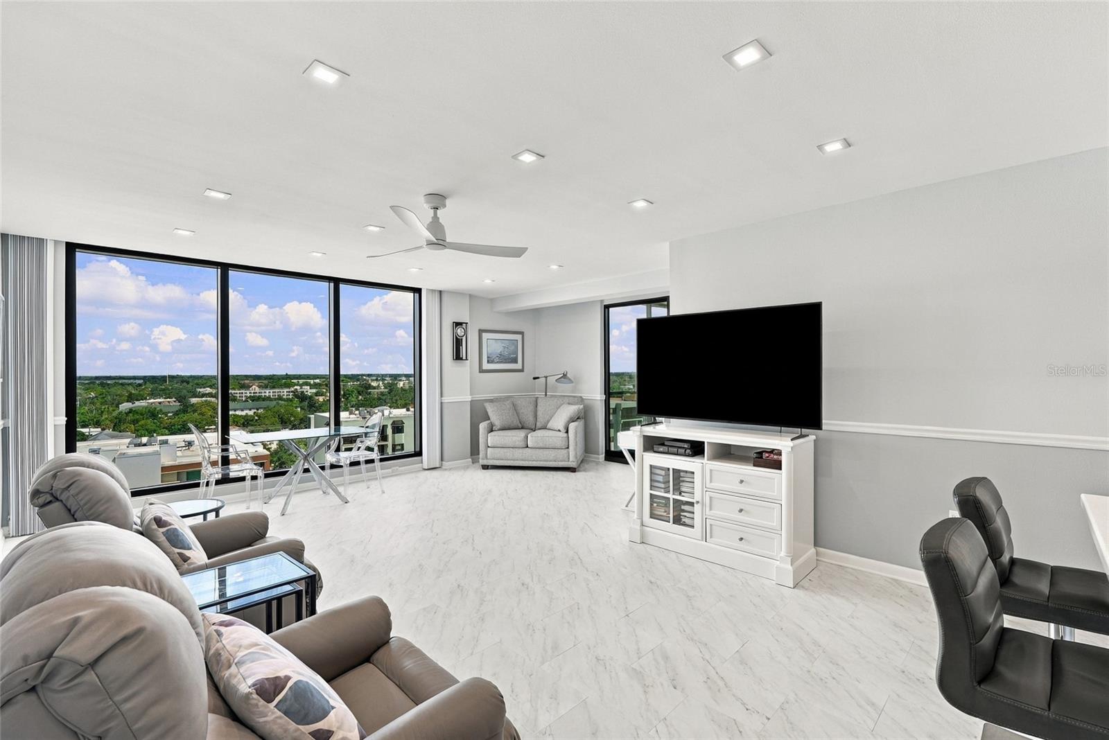 Condominium living room with floor-to-ceiling city-view windows, gray seating, and marble-look floors.