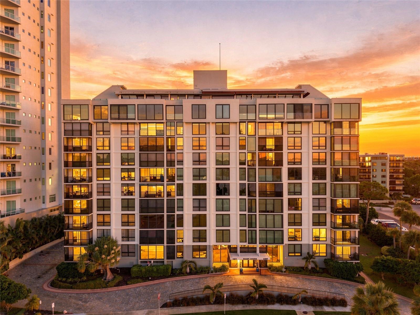 Modern condominium building with glass balconies and illuminated windows at sunset.