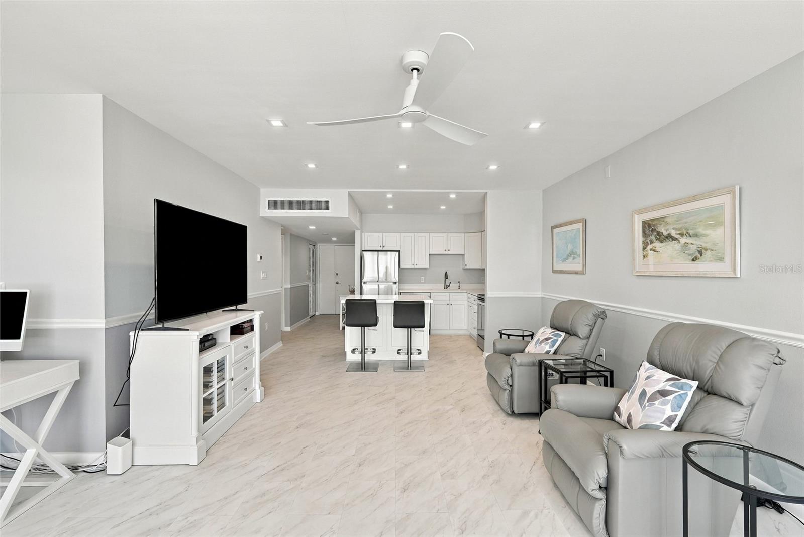Furnished living room with gray recliners, marble-look floors, and an open white kitchen.