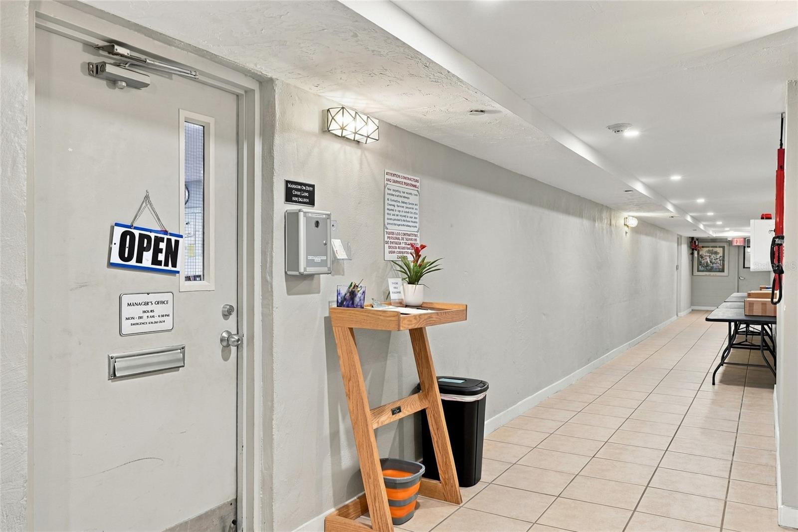 Condominium hallway with a tiled floor, an open manager’s office door, and a wooden console with a potted plant.