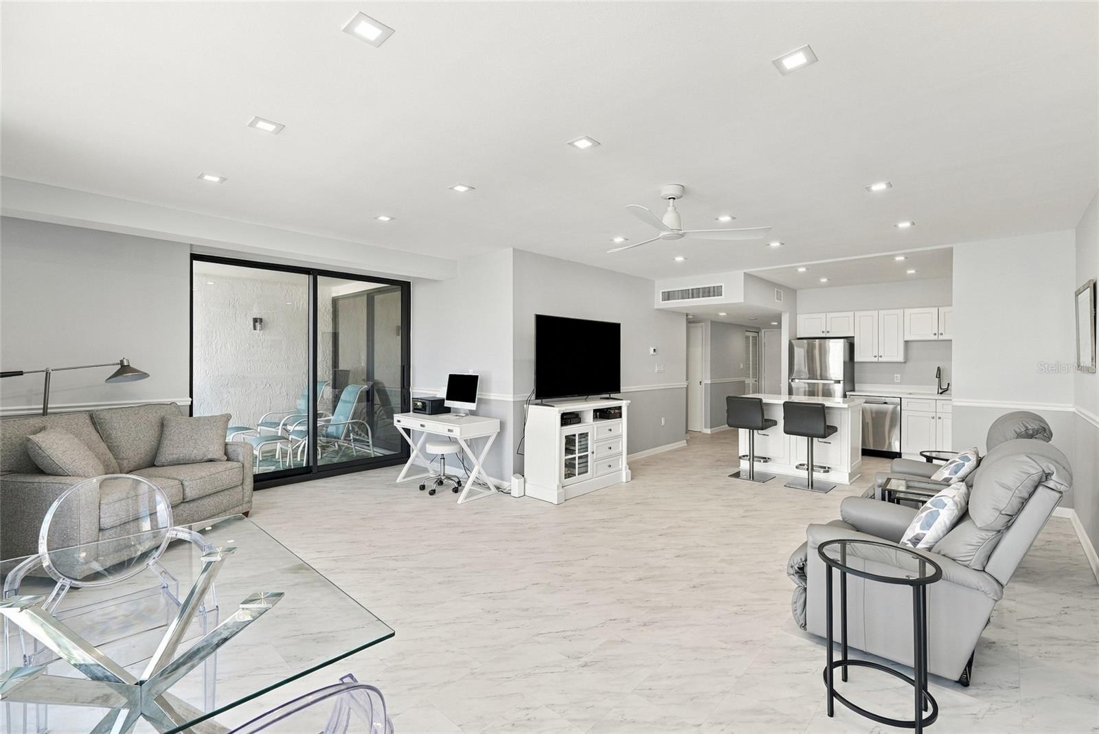 Furnished condo living room with gray seating, marble-look floors, and an open kitchen.