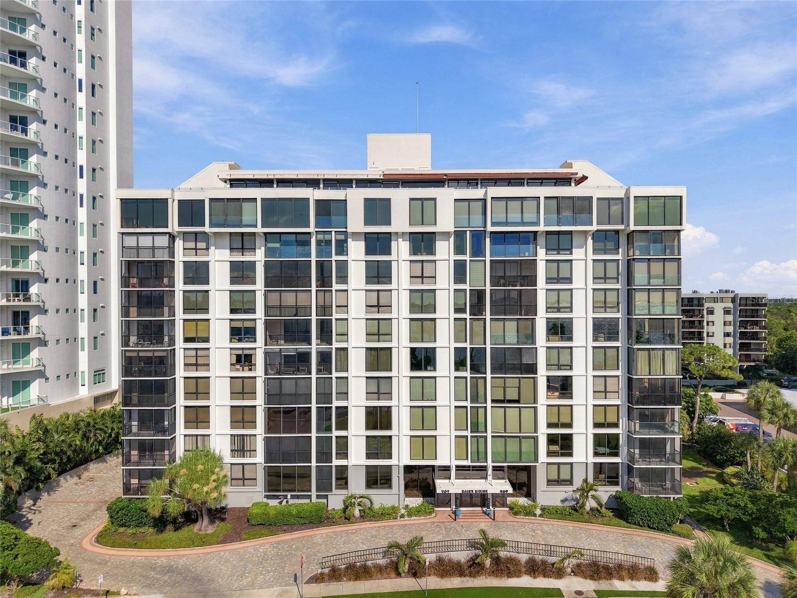 White condominium building with balconies, landscaped entrance, and curved driveway.