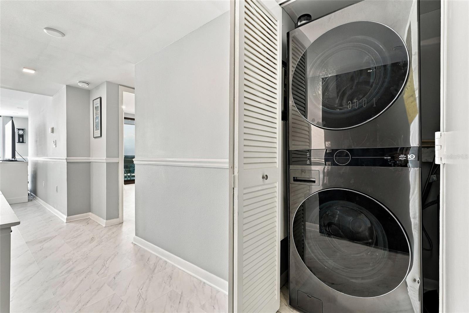 Stacked front-loading washer and dryer in a louvered closet beside a marble-look hallway.