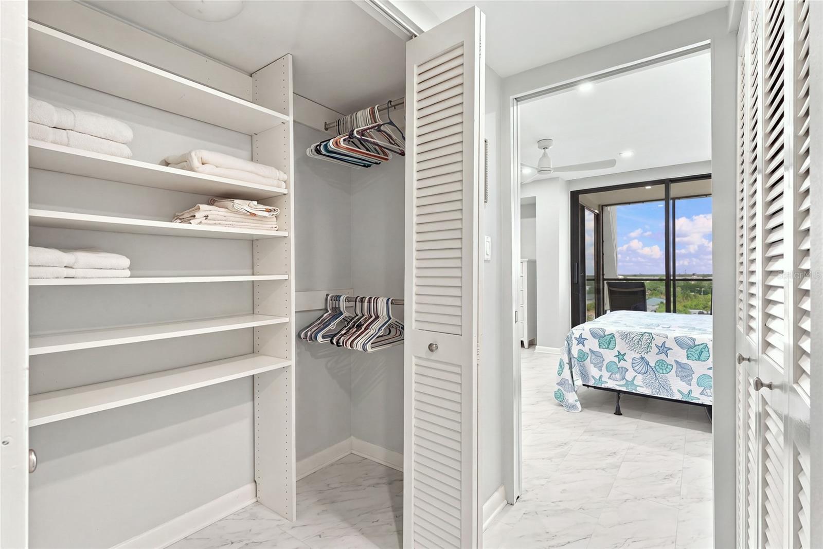 Walk-in closet with built-in shelving and hanging clothes, opening to a bedroom with balcony doors.