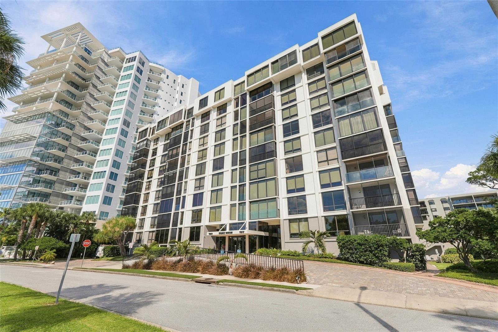 Modern white condominium building with glass balconies and landscaped entrance.