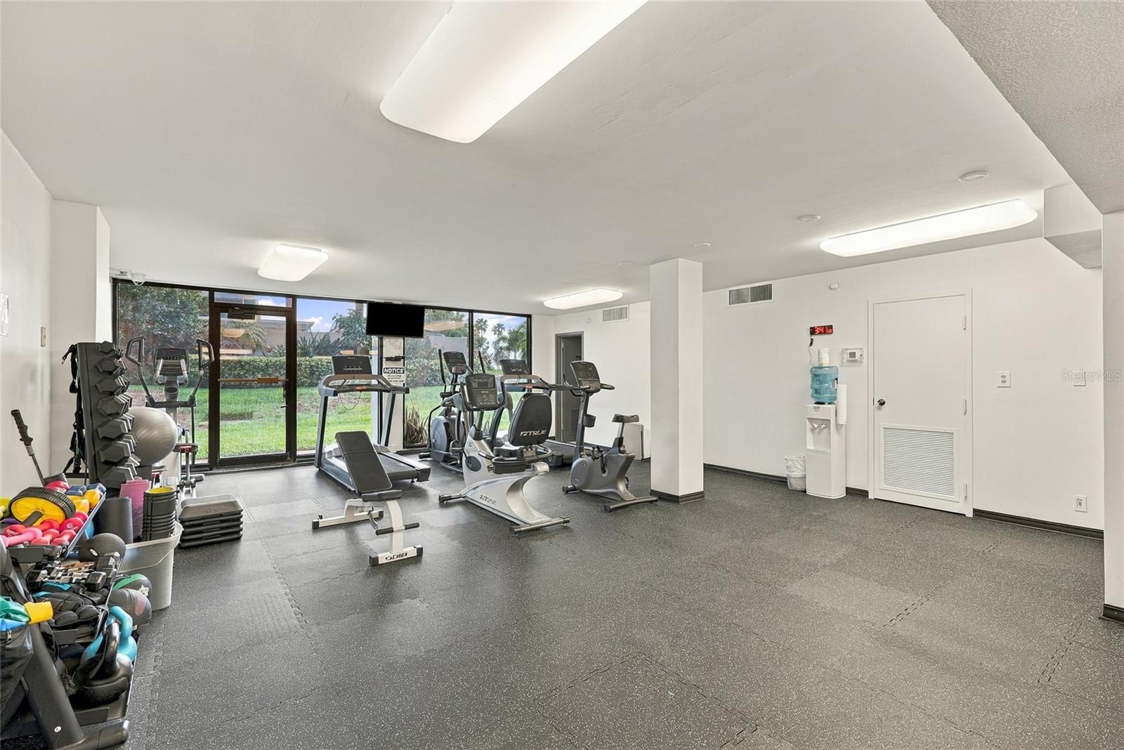 Fitness center with exercise equipment and large windows overlooking a landscaped garden.