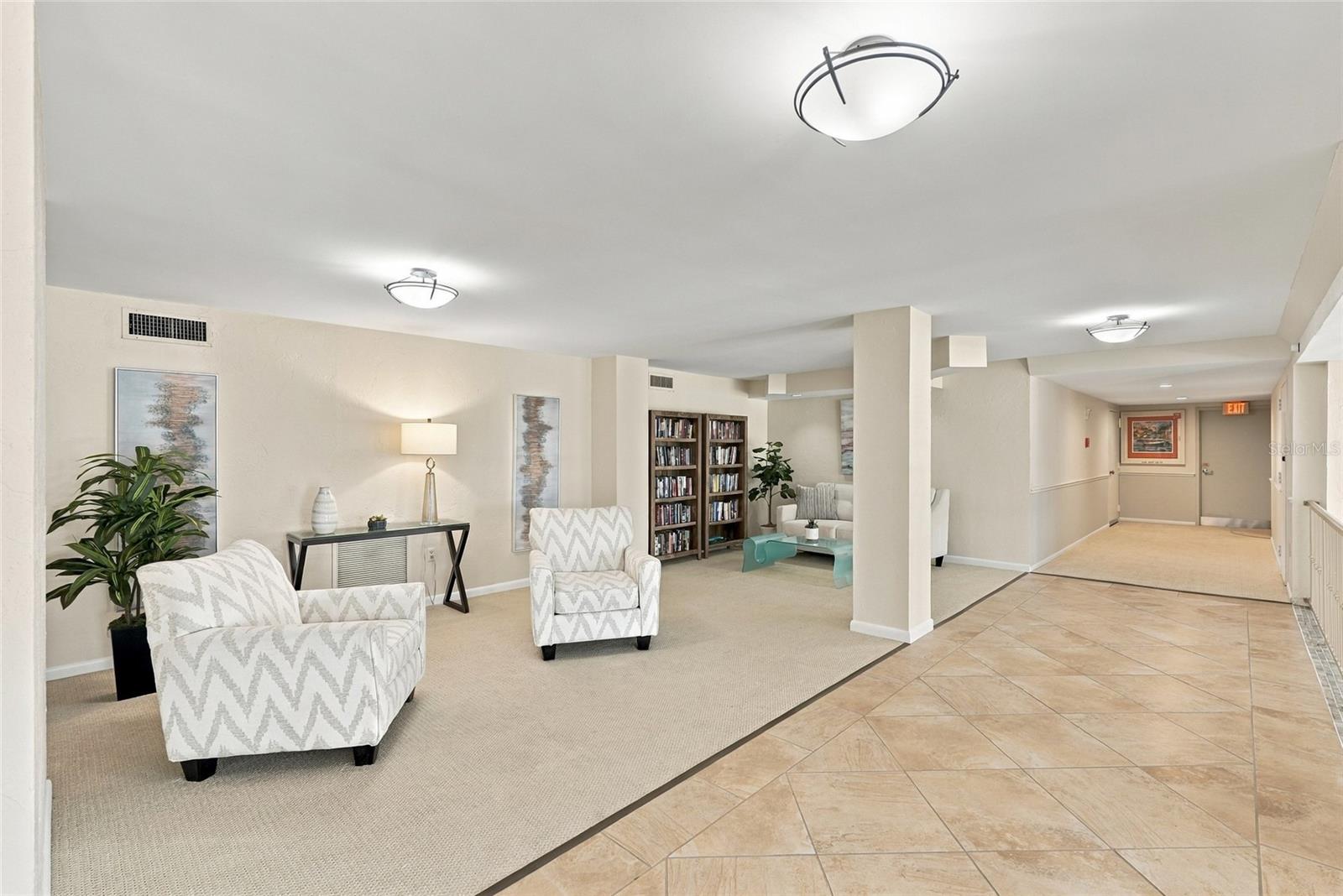 Furnished condominium lobby with patterned armchairs and bookshelves beside a tiled hallway.