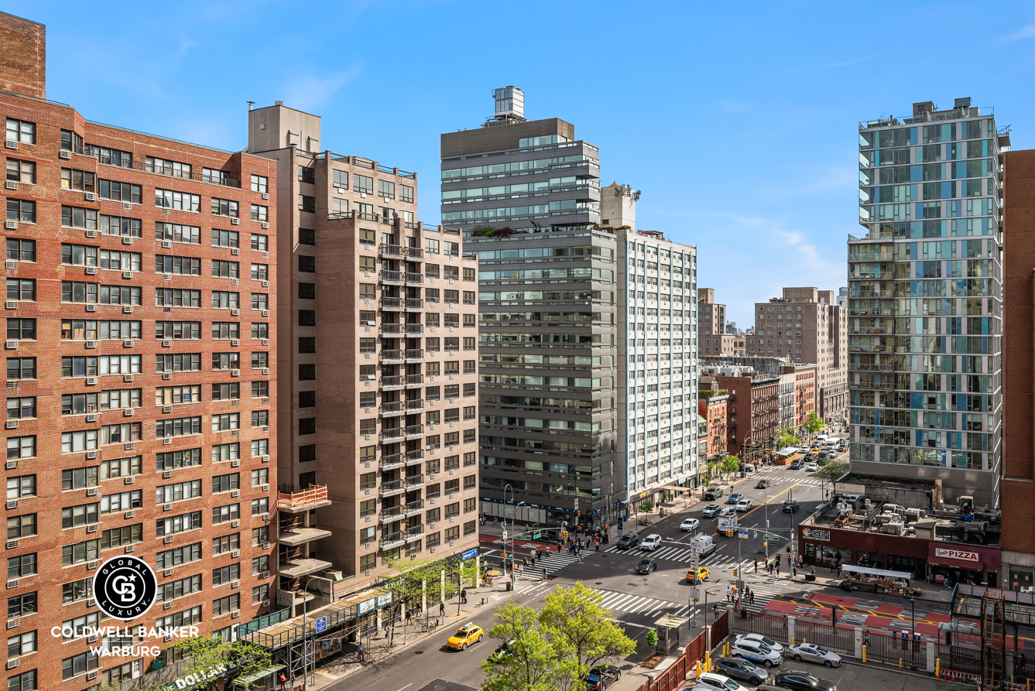 City view over a Manhattan intersection, with mid-rise buildings and a leafy street.