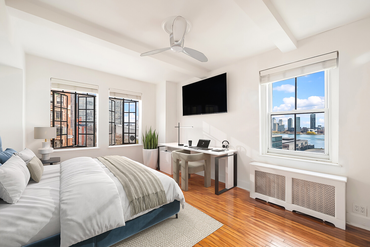 Bright bedroom with hardwood floors, a desk, and windows overlooking the city skyline and water.