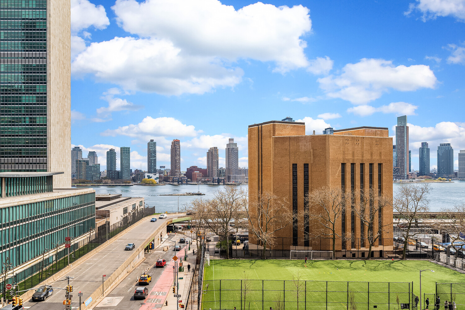 River and city skyline beyond a fenced green sports field and brick building.