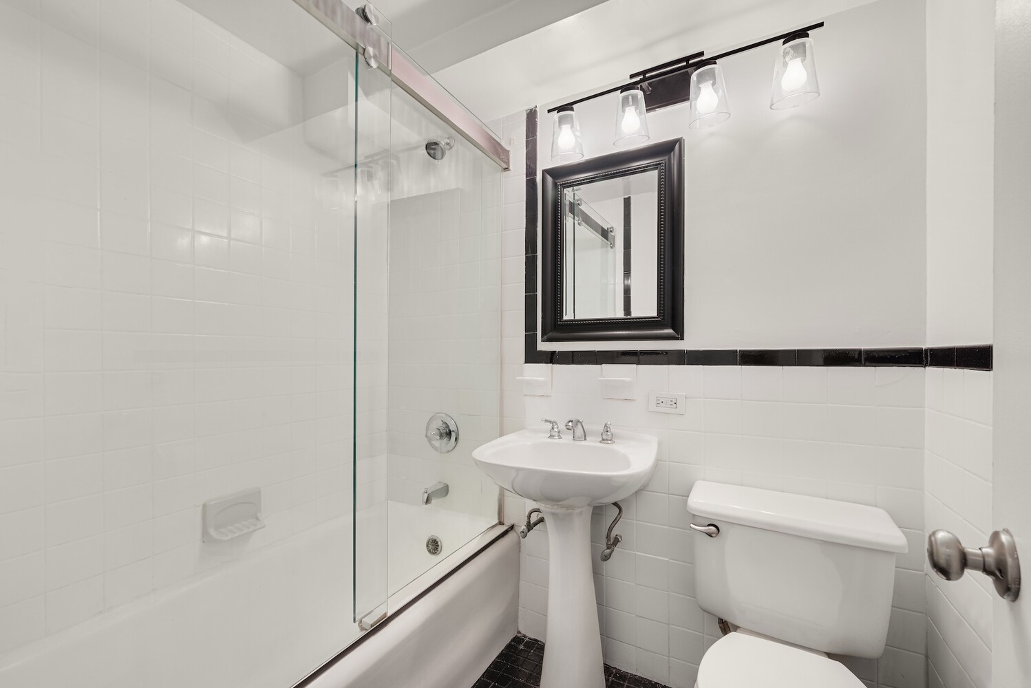 White-tiled bathroom with a glass shower screen, pedestal sink, toilet, and black tile accents.