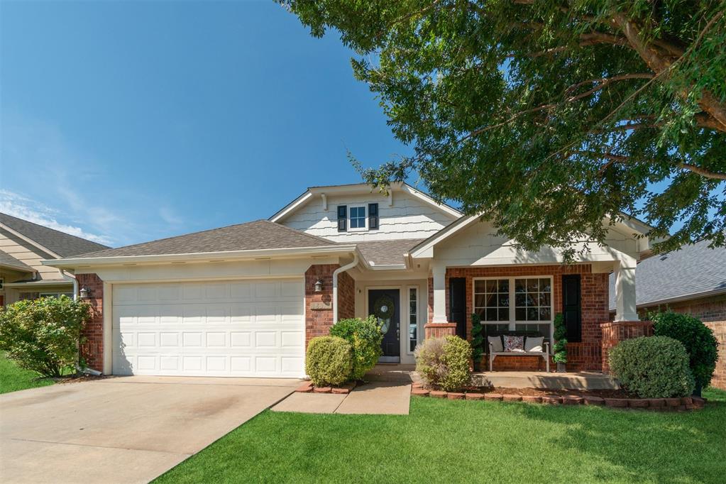 Single-story home with a two-car garage, brick accents, and a covered porch beneath a leafy tree.