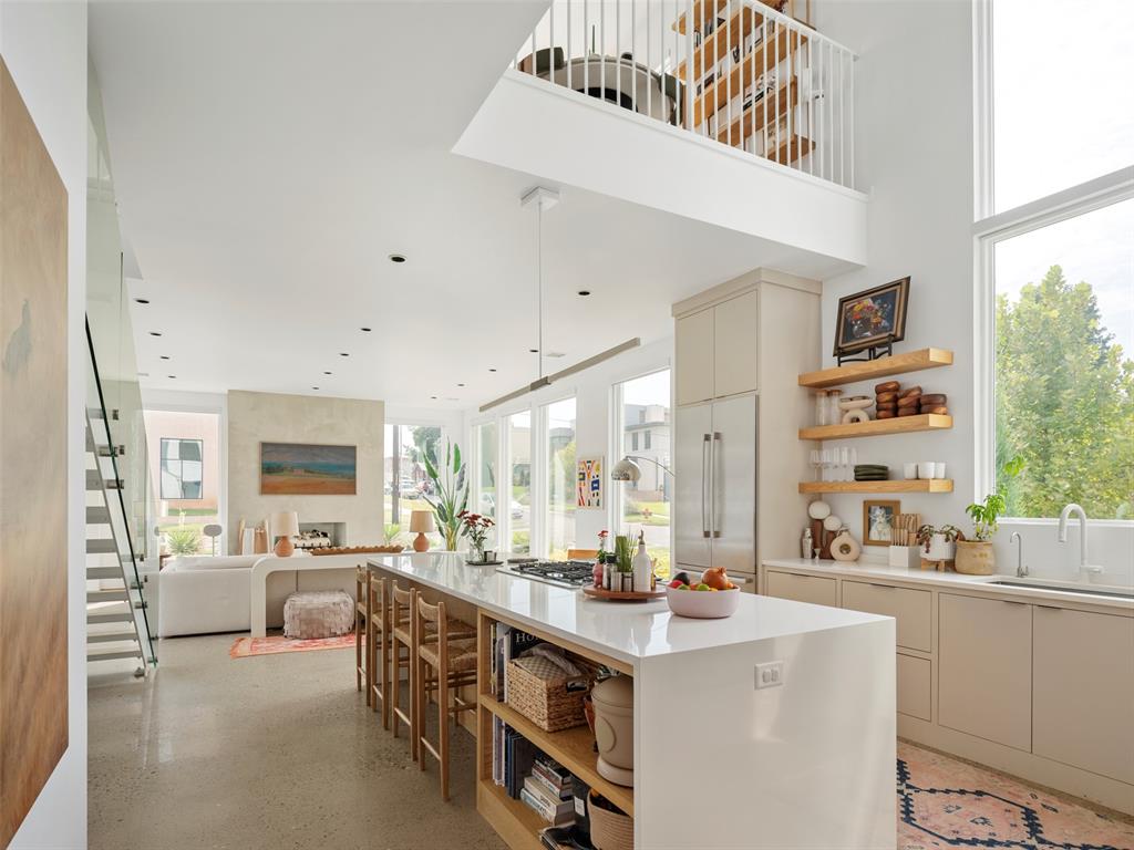 Open-plan kitchen with a long white island, bar stools, large windows, and a staircase.