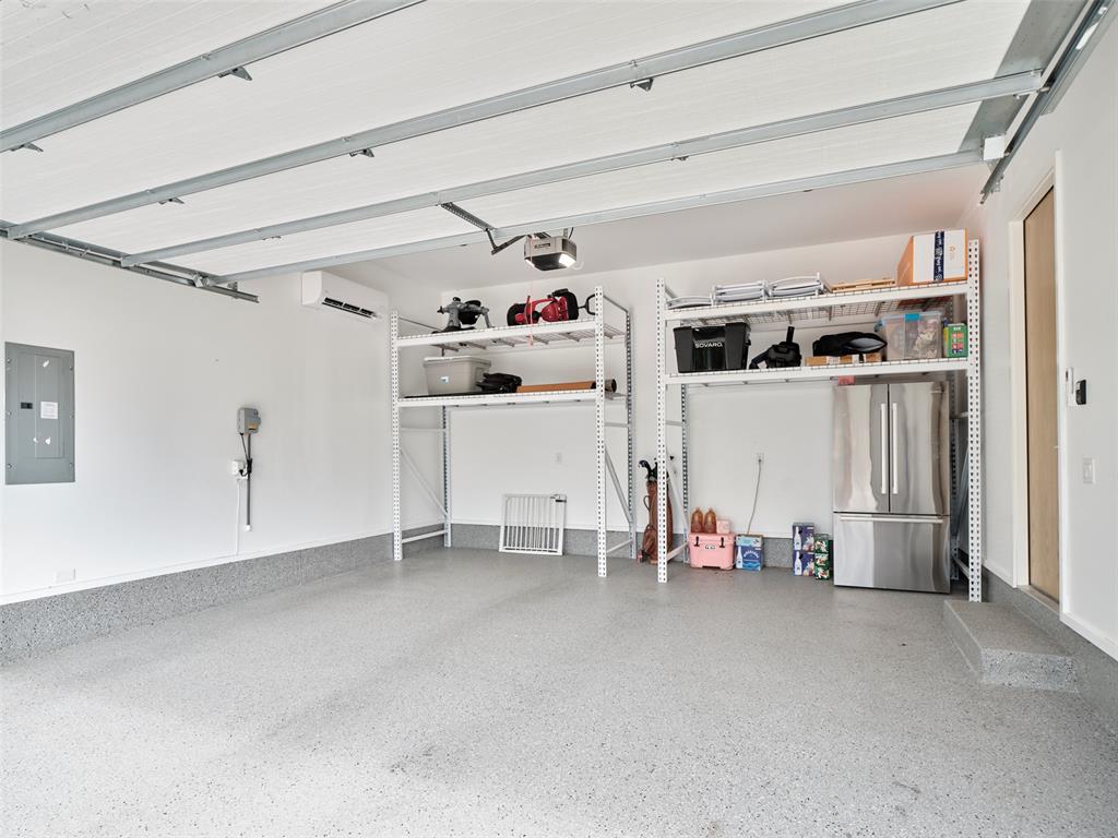 Spacious garage with a speckled floor, open overhead door, storage shelves, and stainless steel refrigerator.