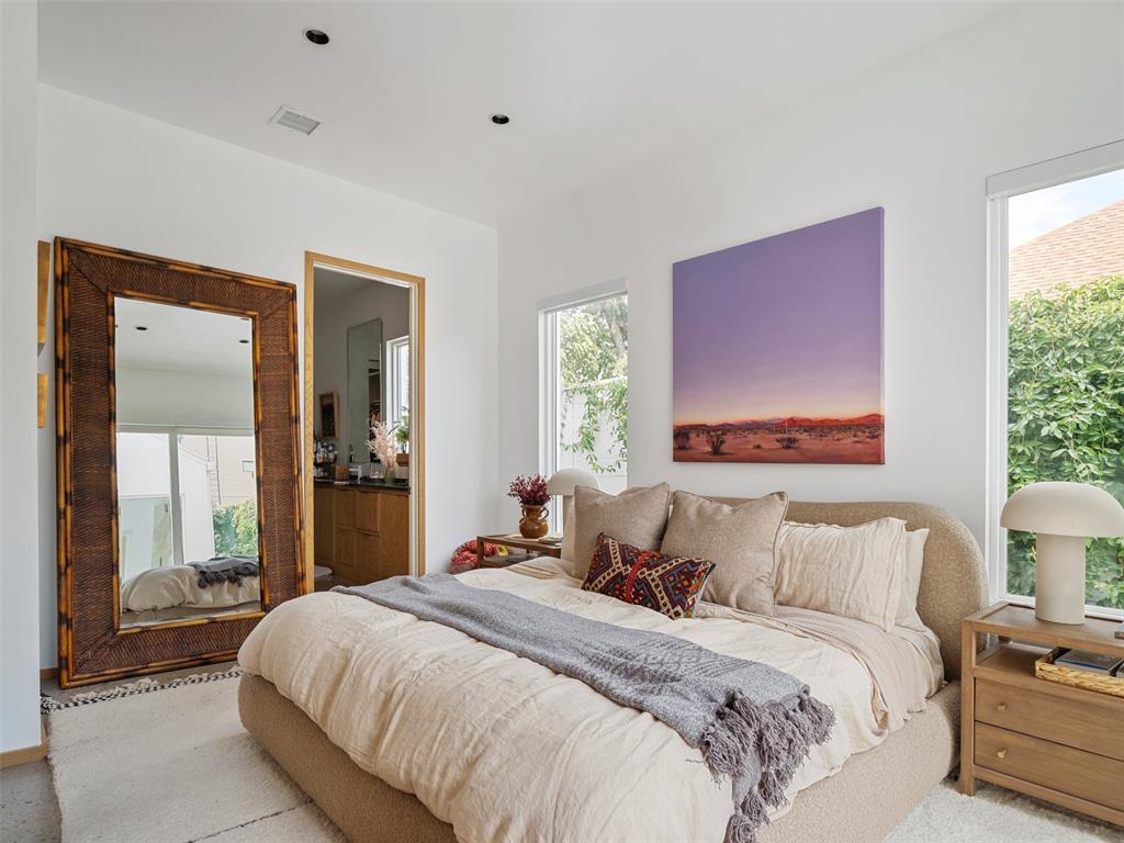 Bright bedroom with a large upholstered bed, woven-frame mirror, and purple landscape artwork.