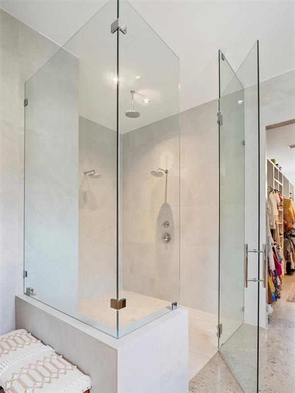 Walk-in shower with frameless glass panels, two showerheads, and pale stone tile.