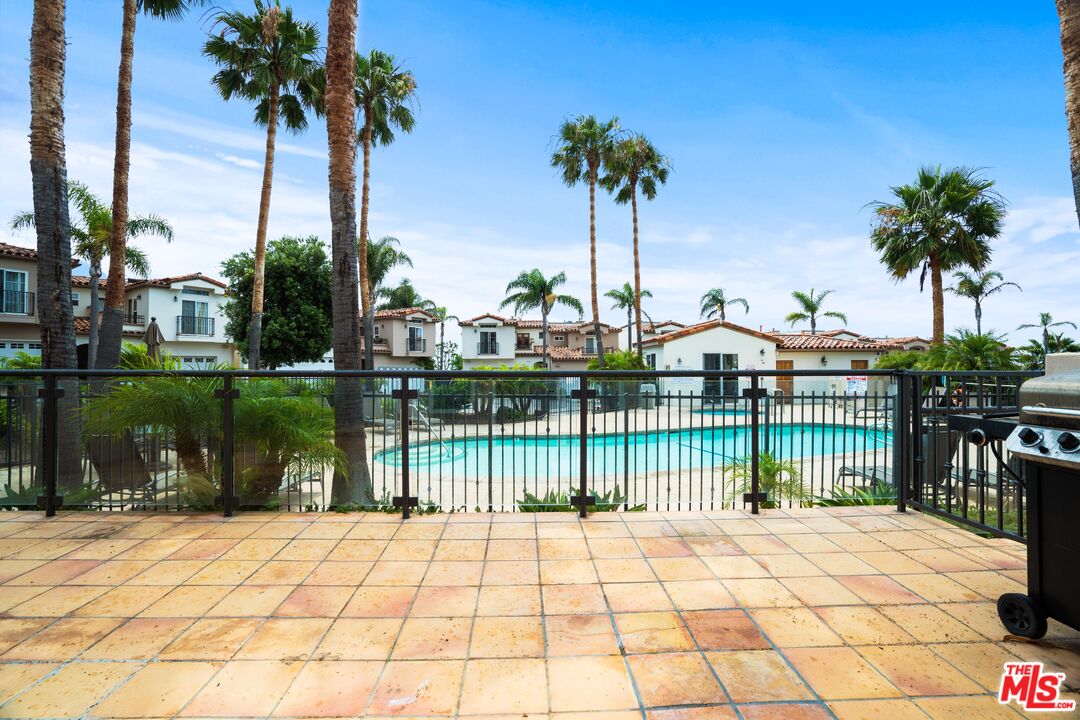 Swimming pool behind a tiled patio and fence, surrounded by palm trees and homes.