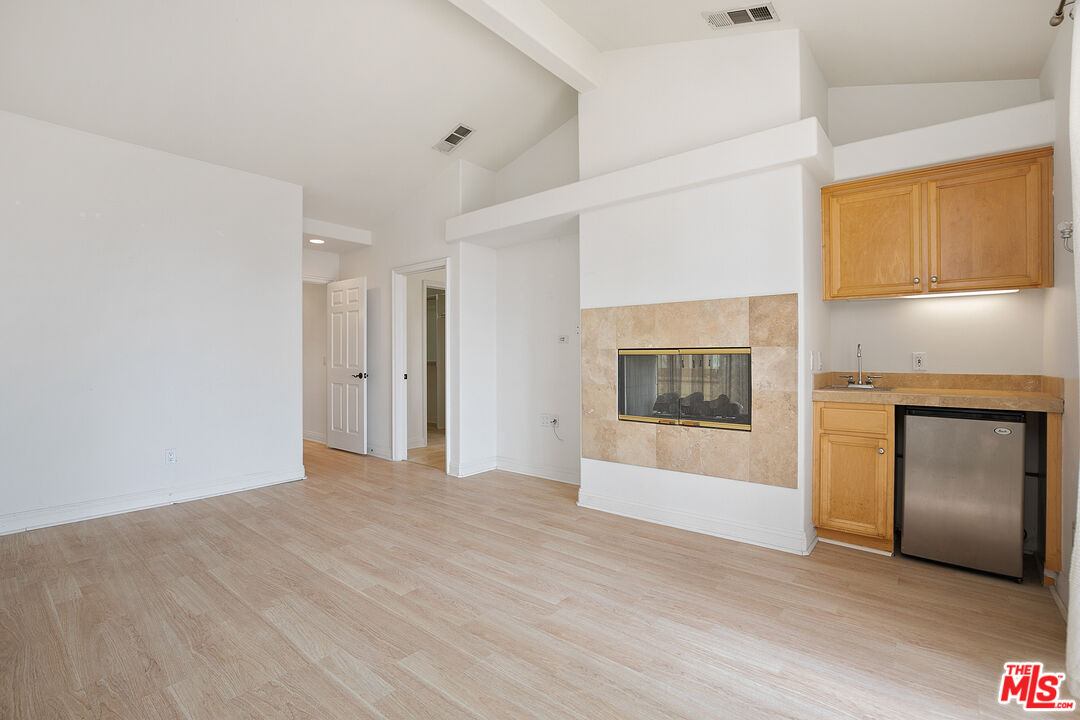 Vacant room with a tiled fireplace, light wood floors, and a kitchenette with wood cabinets.