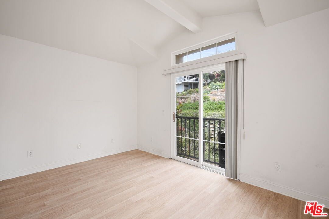 Empty bright room with pale wood floors, vaulted ceiling, and sliding doors to a balcony.