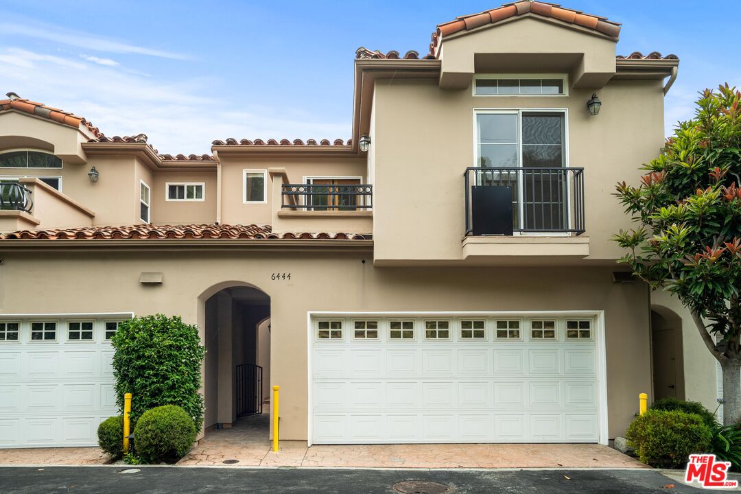 Two-story beige home with a tiled roof, arched entry, balcony, and white double garage door.