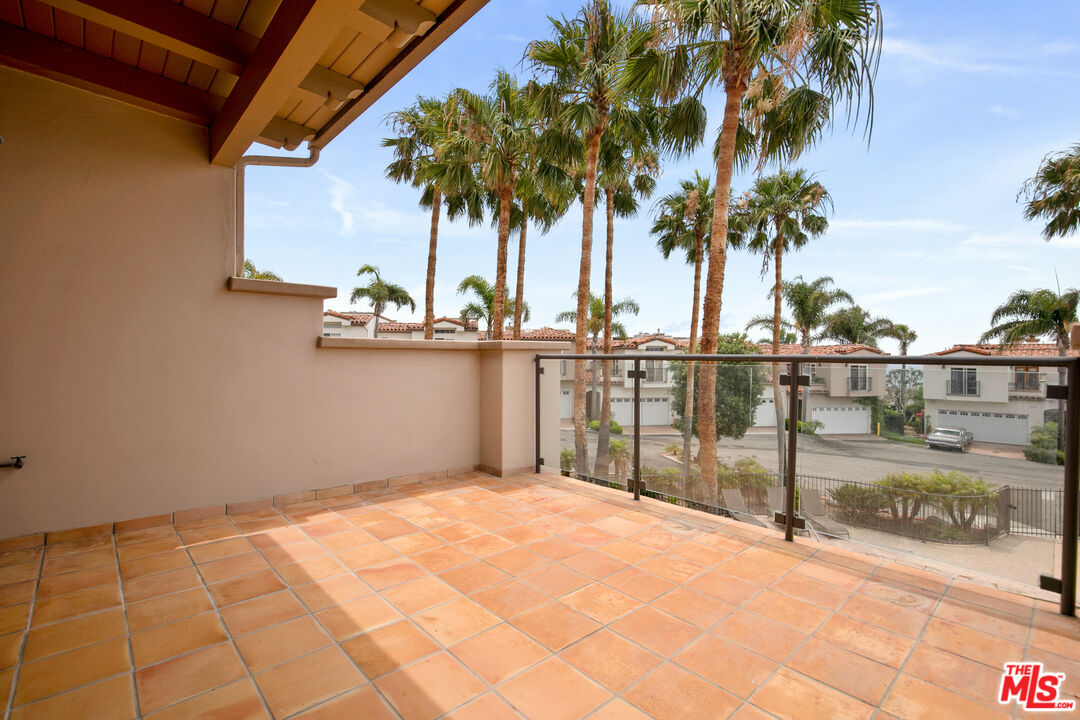 Covered tiled balcony with glass railing overlooking palm trees and neighboring homes.