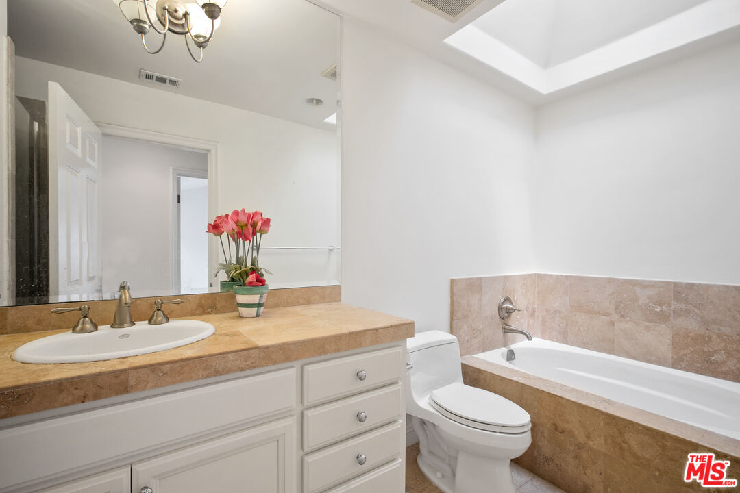 Bathroom with a tan stone vanity and bathtub, large mirror, and pink flowers on the counter.