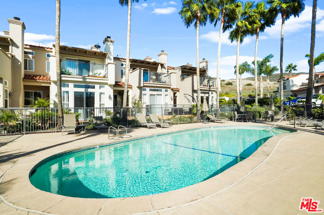 Outdoor swimming pool with turquoise water, lounge chairs, and palm trees in front of beige homes.