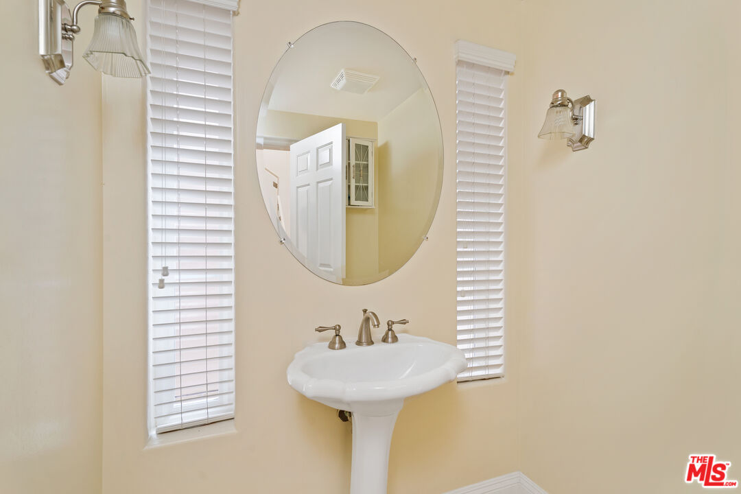 Bathroom pedestal sink beneath an oval mirror, flanked by narrow windows with white blinds.