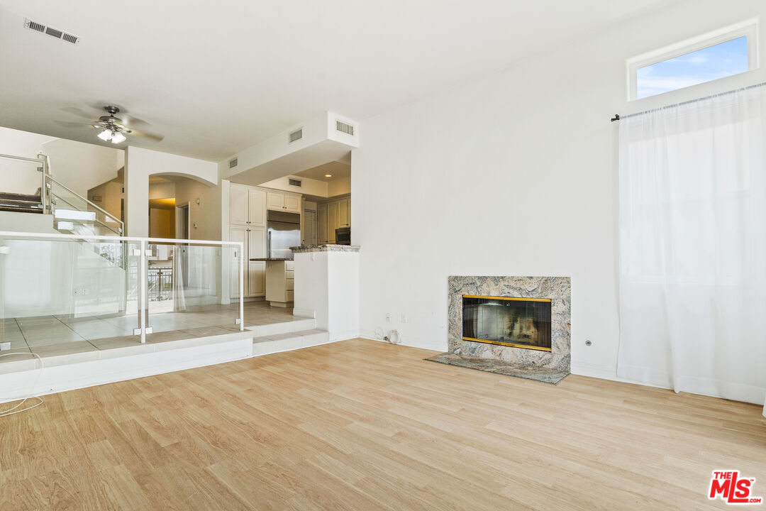 Vacant living room with light wood floors, stone fireplace, and glass railing by the kitchen.