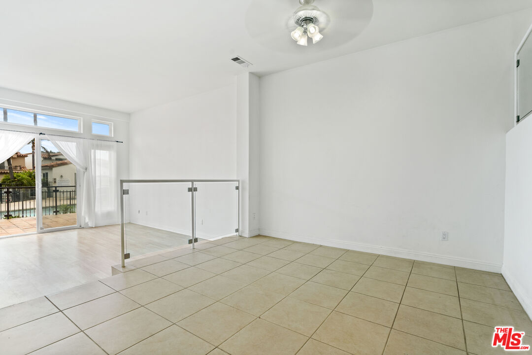 Vacant tiled room with a glass railing and sliding doors opening to a balcony overlooking a pool.