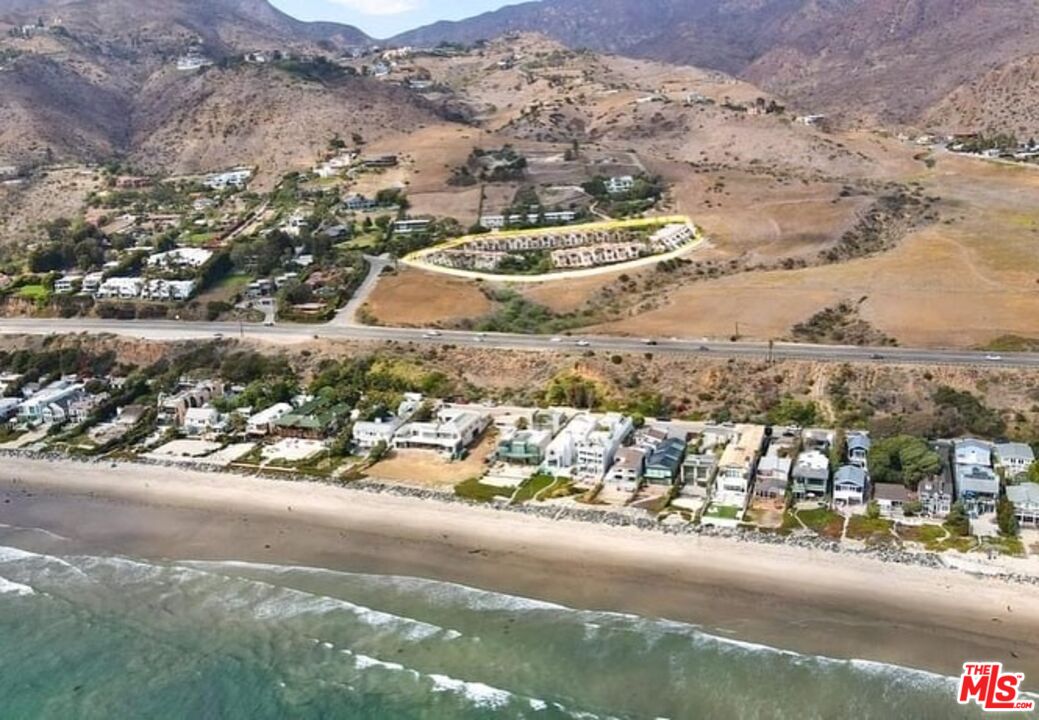 Aerial view of Malibu beachfront homes, with waves, a coastal road, and dry mountains inland.