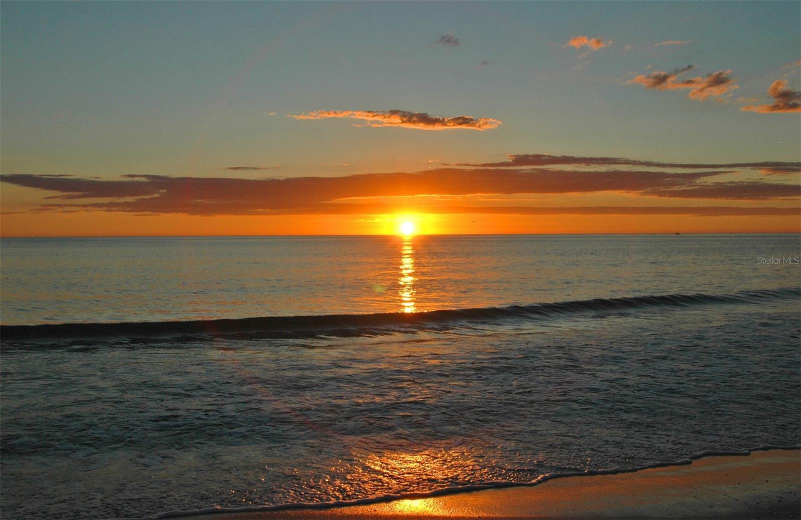 Sunset over the ocean, with orange light reflecting on the water beside a sandy beach.