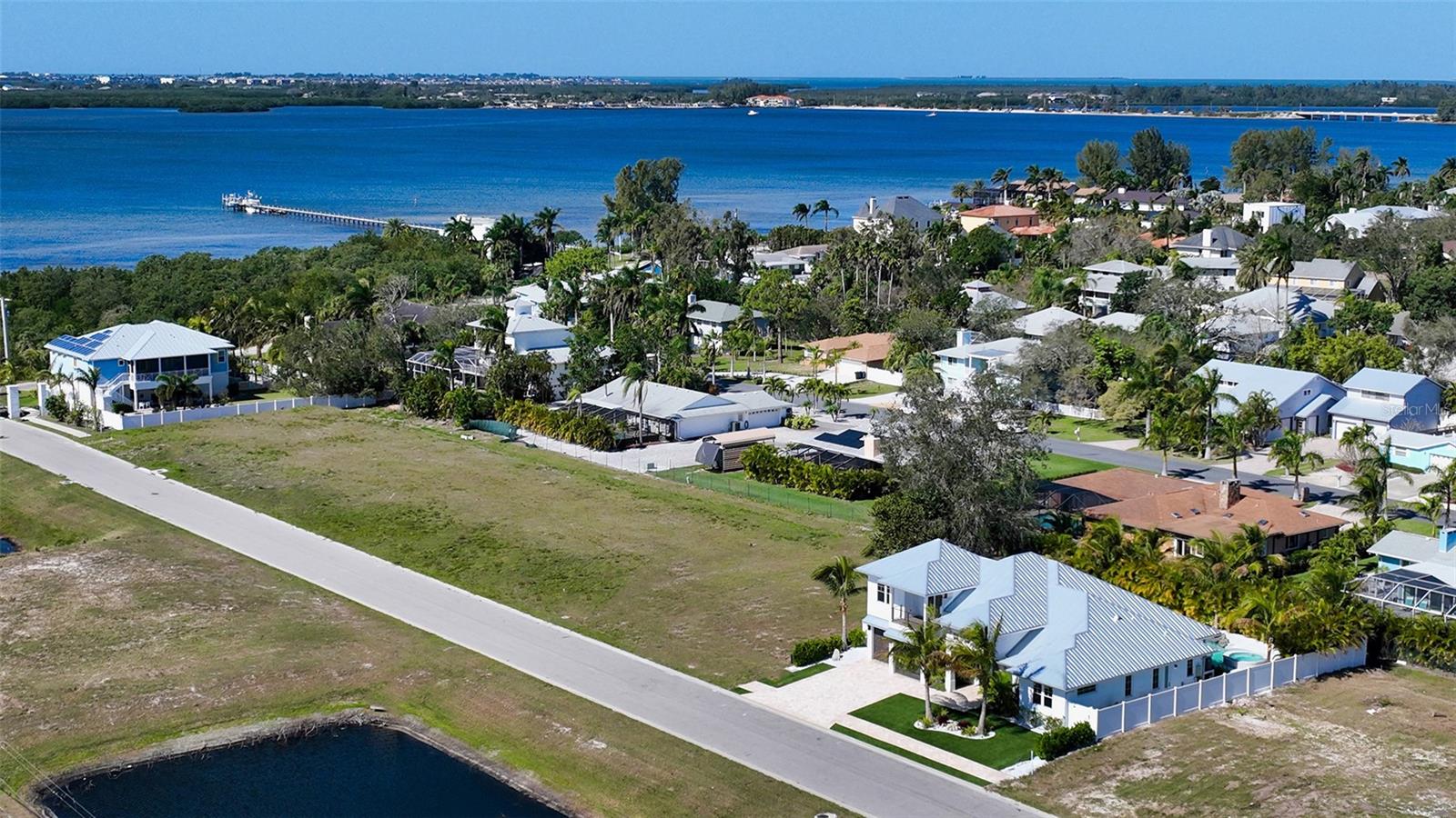 Vacant grassy lot beside a road, with coastal homes, a pier, and the bay beyond.