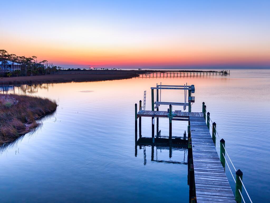 A wooden dock extends into calm bay water beneath a colorful sunset.