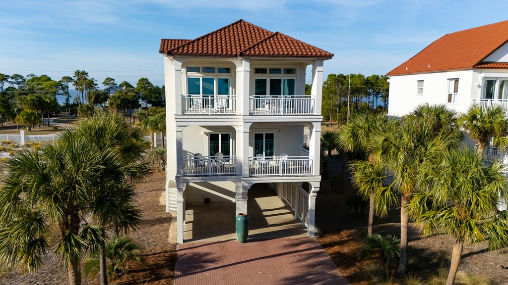 White two-story coastal home with red tile roof, stacked balconies, and covered parking, framed by palm trees.