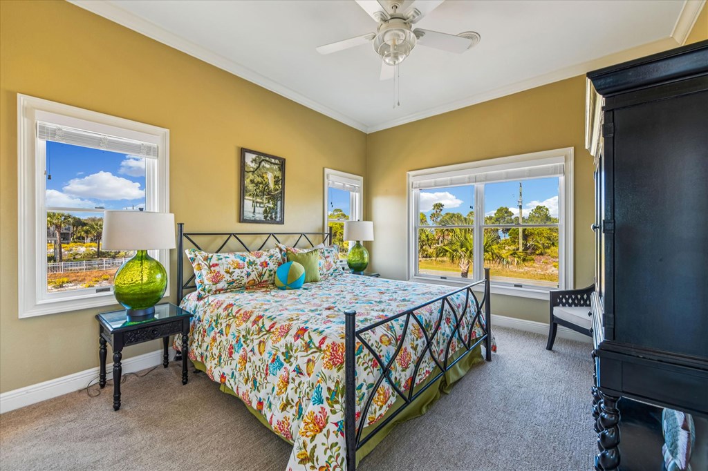 Furnished bedroom with colorful floral bedding, mustard-yellow walls, and windows overlooking palm trees.