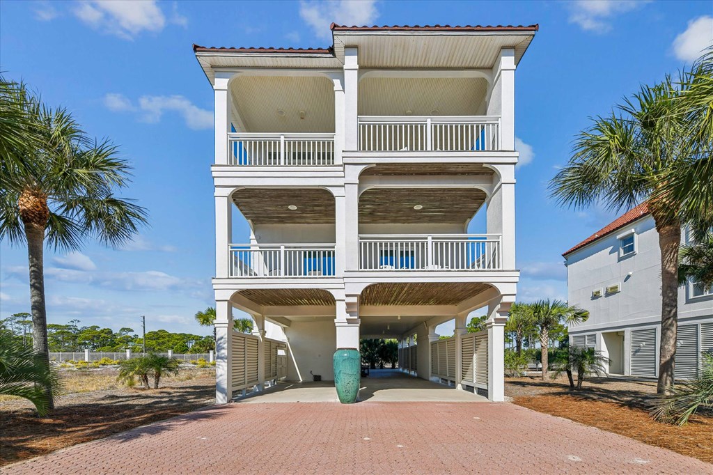 White elevated coastal house with three covered balconies, a ground-level carport, and a brick-paved driveway.