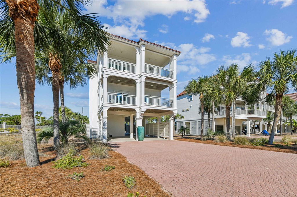 White coastal house with stacked covered balconies, palm trees, and a wide brick-paved driveway.