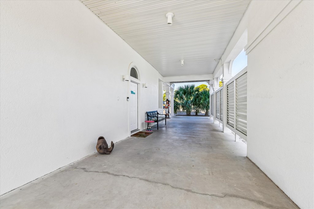 Covered concrete porch with a white arched door, bench, louvered shutters, and palms at the far end.